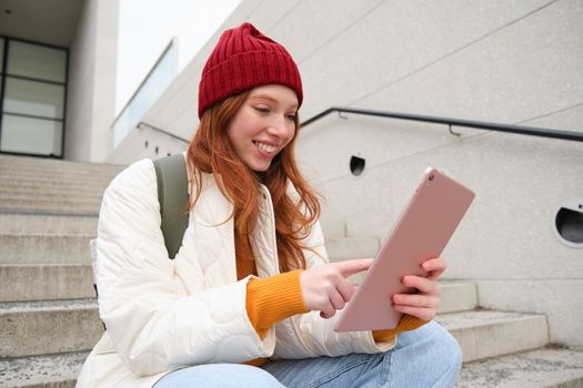 Happy stylish redhead girl, student in red hat, holds digital tablet, uses social media app, searches something online, connects to wifi.