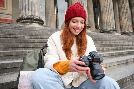 Young student, photographer sits on street stairs and checks her shots on professional camera, taking photos outdoors.