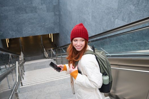 Smiling girl walking on stairs, using mobile phone, texting message on her way, using smartphone app map, follows route to destination place.