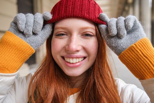 Headshot of happy redhead girl with freckles, wears red hat and gloves in winter, walks around city on chilly weather and smiles.