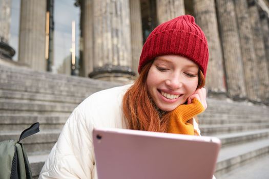 Portrait of young redhead woman sitting outdoors on stairs, reading e-book on digital tablet, wearing red hat and warm clothes.