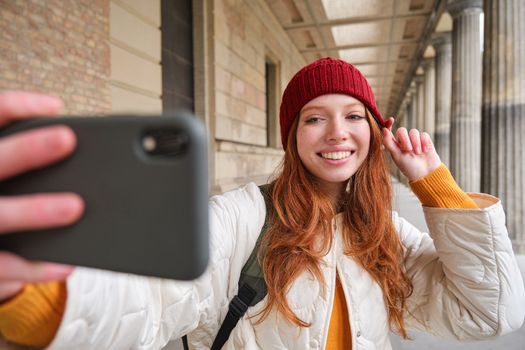 Cute young redhead woman takes selfie on street with mobile phone, makes a photo of herself with smartphone app on street.