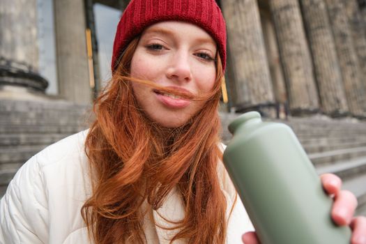 Portrait of redhead woman drinking from thermos, sitting on street stairs and enjoys hot drink from flask.