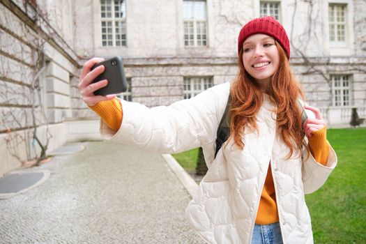 Portrait of happy girl tourist, takes selfie on smartphone in front of historical building, posing for photo on mobile phone camera.