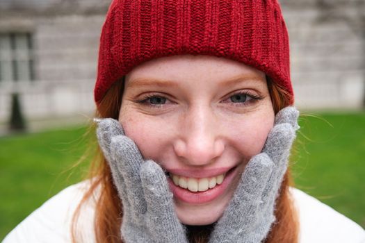 Close up portrait of beautiful redhead woman in red knitted hat, warm gloves, smiling and looking happy at camera, sitting in park.