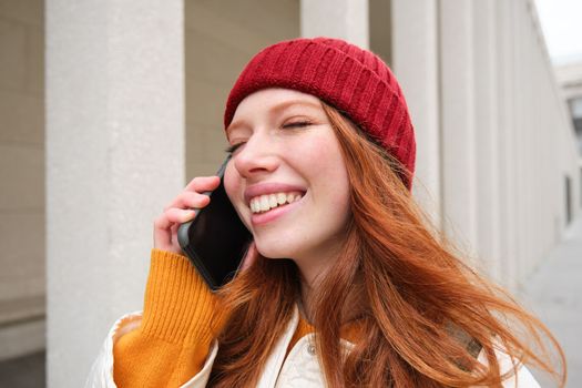 Mobile broadband and people. Smiling young redhead woman walks in town and talks on mobile phone, calling friend on smartphone, using internet to make a call abroad.