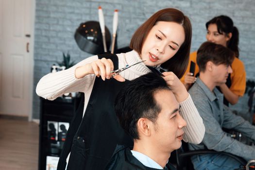 Asian female hairdresser making haircut for two male customer with male hairstyle in qualified barbershop. Men's hairstyling by a scissor and comb in hair salon concept.