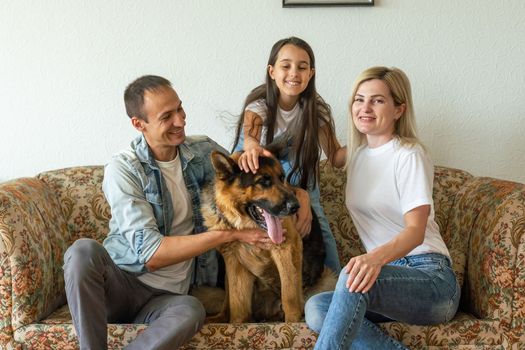 Beautiful young parents, their cute little daughter looking at camera and smiling, sitting with their cute dog on sofa at home.