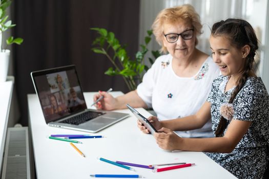 Portrait of happy grandmother and little granddaughter making video conference on pc sitting at table, waving hands at screen, greeting somebody, chatting with parents, enjoying online communication