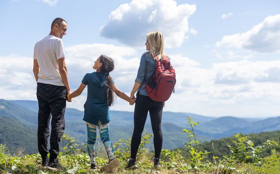 Family standing with arms around on top of mountain, looking at beautiful summer mountain landscape. People enjoying view