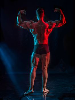 Handsome young bodybuilder doing classic double biceps pose, looking away, on greenish background, full figure shot