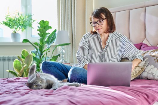 Middle aged woman at home on bed with laptop and cat. Mature 40s female looking at screen, on sofa with pet. Work at home, technology, leisure, freelancing, lifestyle, people and animals concept