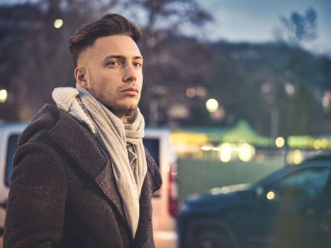 Handsome trendy young man, standing on a sidewalk in city setting at night wearing a fashionable winter coat and scarf, looking at camera