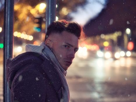 Handsome trendy young man, standing on a sidewalk in city setting at night wearing a fashionable winter coat and scarf