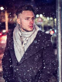 Handsome trendy young man, standing on a sidewalk in city setting at night wearing a fashionable winter coat and scarf