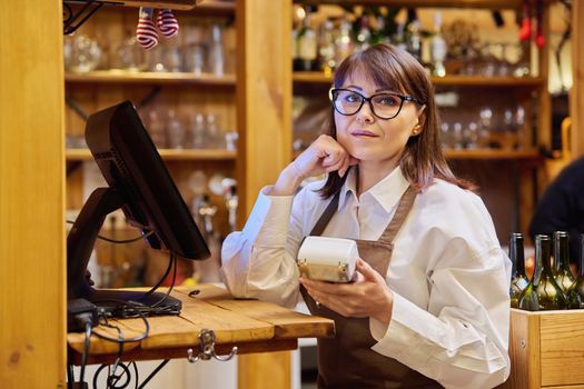 Portrait of woman in apron worker manager bar restaurant looking at camera with wireless bank terminal for processing credit card payments. Cashless technology money service work small business staff