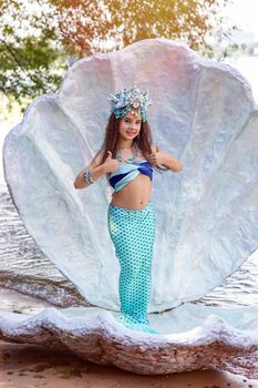 A beautiful little girl in a mermaid costume stands outdoor, in a large sea shell, holds her thumbs up. Copy space. Vertical