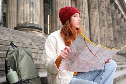 Beautiful young woman, traveler with backpack holds paper map, explores city sightseeing, plans route for tourism popular attractions.