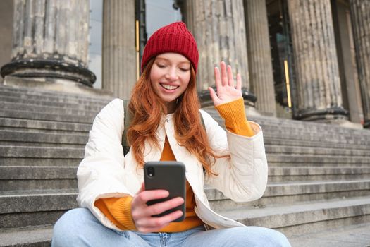 Young redhead woman sits on stairs outdoors and waves hand at smartphone camera, video chats with friends, connects to public wifi.