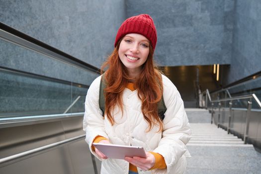 Beautiful redhead female model posing in city, walking up stairs with digital tablet, using gadget to plan her route, reading while going somewere.