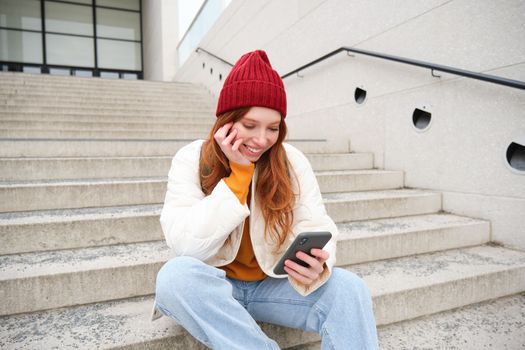 Beautiful redhead teen girl, sits on stairs and smiles, holds mobile phone, uses smartphone application, chats on telephone app.