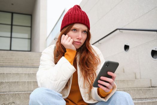 Portrait of sad redhead girl, looks upset and disappointed at smartphone screen, reads bad news text message on mobile phone and frowns, sits on stairs outdoors.