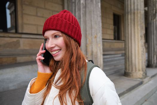 Smiling young redhead woman listens to voice message, makes a phone call, walks on street and talks to someone on smartphone.