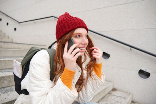 Beautiful smiling redhead female model, sits on street and talks on mobile phone, uses smartphone app to call abroad, laughing during telephone conversation.