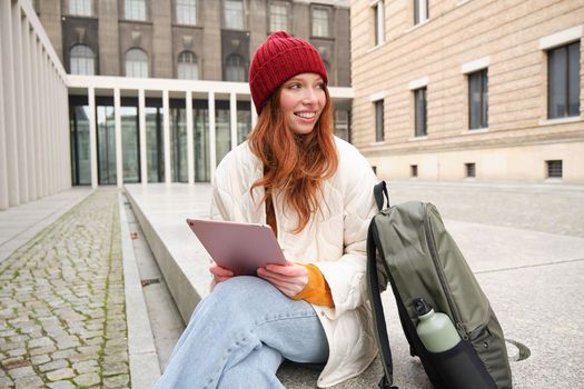 Redhead girl smiles, sits outdoors near building with digital tablet, thermos and backpack, connects to public internet and searches smth online on her gadget.