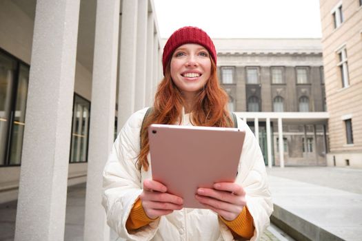 Happy redhead girl in red hat, walks around city with digital tablet, connects to public internet wifi and looks for route, looks at map on her gadget.