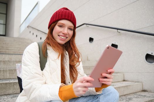 Happy stylish redhead girl, student in red hat, holds digital tablet, uses social media app, searches something online, connects to wifi.