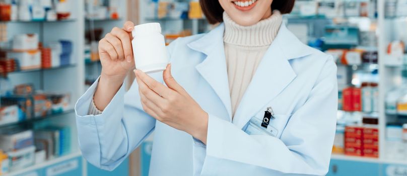 Closeup young affable pharmacist show qualified medicine pill bottle mockup product for copyspace at pharmacy. Druggist working with her diary job at drugstore, medicine box on shelf at background