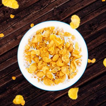 Bowl of sweet cornflakes with milk dark wooden table background, top view. Corn flakes with milk splashes dark wooden table