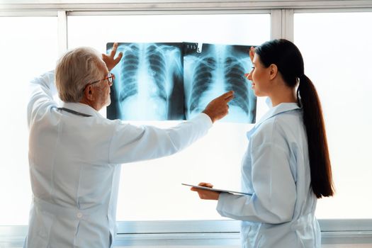 In a hospital sterile room, two professional radiographers hold and examine a radiograph for medical xray diagnosis. Novice doctor seeks advice on a patient's condition from experienced older doctor.