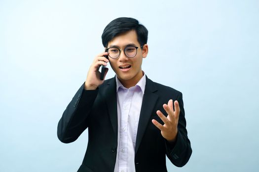 Portrait of young smart asian businessman in eyeglasses standing, smiling, and talking on mobile phone about business with light blue isolated background. Business and connection concept.