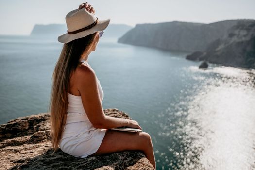 Successful business woman in yellow hat working on laptop by the sea. Pretty lady typing on computer at summer day outdoors. Freelance, travel and holidays concept.