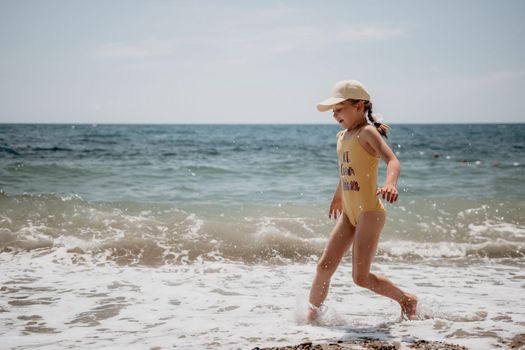 Cute little girl running along the seashore against a clear blue sea and rejoices in the rays of the summer sun. Beautiful girl in yellow swimsuit running and having fun on tropical beach