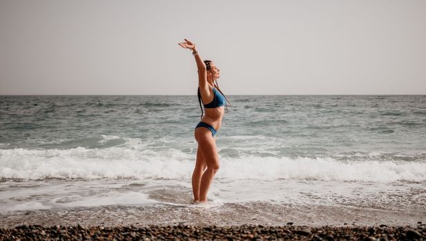 Beach vacation. Hot beautiful woman in sunhat and bikini standing with her arms raised to her head enjoying looking view of beach ocean on hot summer day.