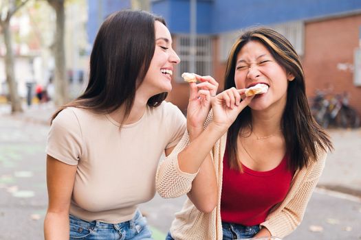 couple of young women sharing some sweet buns and playing jokes sitting in a city park, concept of friendship and love between people of the same sex