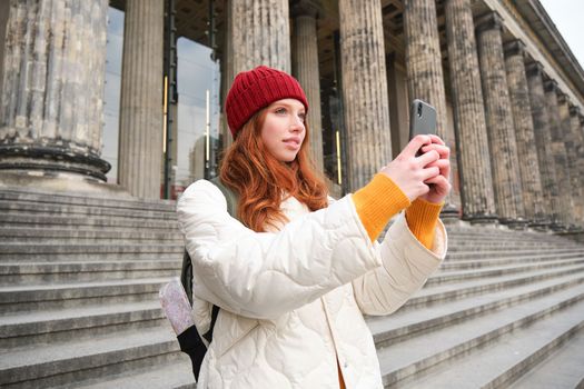 Portrait of young blogger, redhead girl tourist takes pictures of sightseeing, makes photos on smartphone camera, records video and smiles.