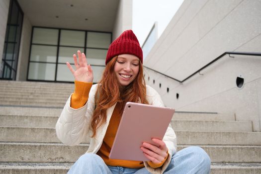 Joyful redhead teen girl, student with digital tablet, says hello, waves hand at gadget camera, connects to video chat, talks to friend in application.