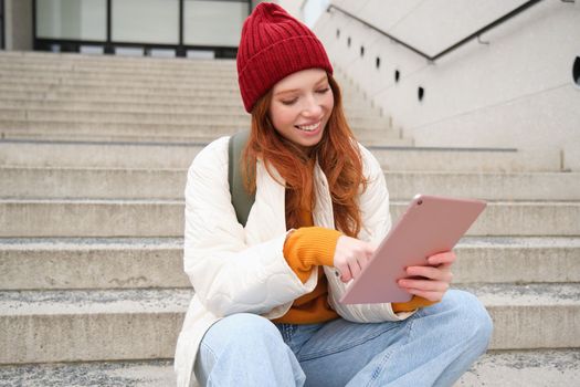 Happy stylish redhead girl, student in red hat, holds digital tablet, uses social media app, searches something online, connects to wifi.