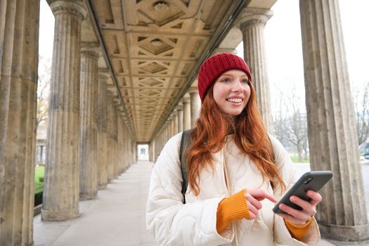 Happy young female model stands on street and holds mobile phone, uses smartphone app, follows the route of online map.