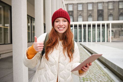 Happy redhead girl in red hat, walks around city with digital tablet, connects to public internet wifi and looks for route, looks at map on her gadget.