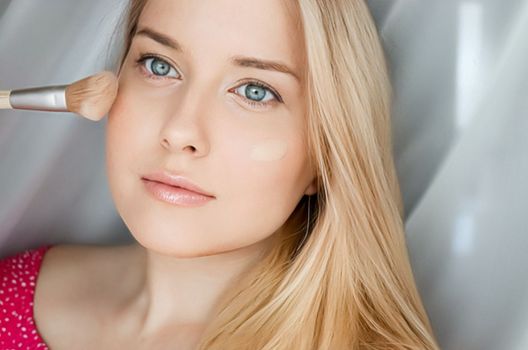 Beautiful blonde woman applying liquid make-up foundation on her skin with make-up brush.