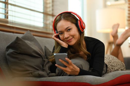 Satisfied millennial woman with wireless headphone lying on comfortable couch and using smart phone.