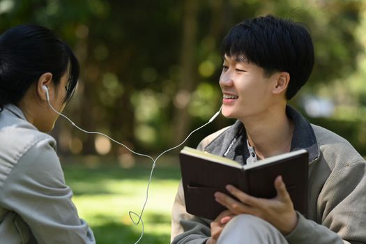 Smiling young asian man student reading a book and listening to music in earphones with his girlfriend.