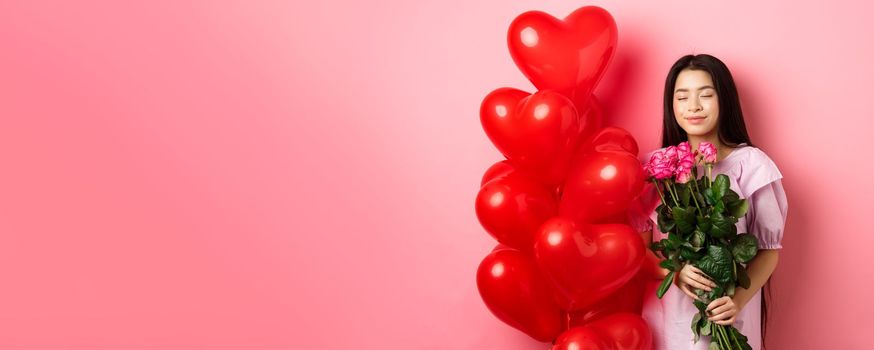 Valentines day concept. Romantic teen asian girl dreaming of love or date, close eyes and smile, holding flowers from lover, receive bouquet of roses and red heart balloons, pink background.