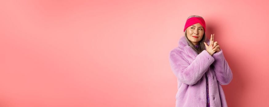 Fashion and shopping concept. Cool and stylish asian senior lady in purple fake fur coat, making finger gun gesture and looking left with sassy smile, acting like secret agent, pink background.