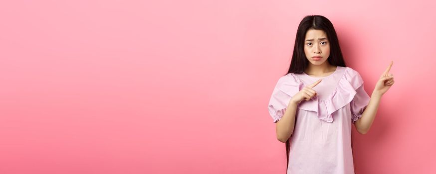 Sad and distressed asian girl frowning, sulking and pointing fingers right at disappointing bad news, feeling unhappy, standing against pink background.
